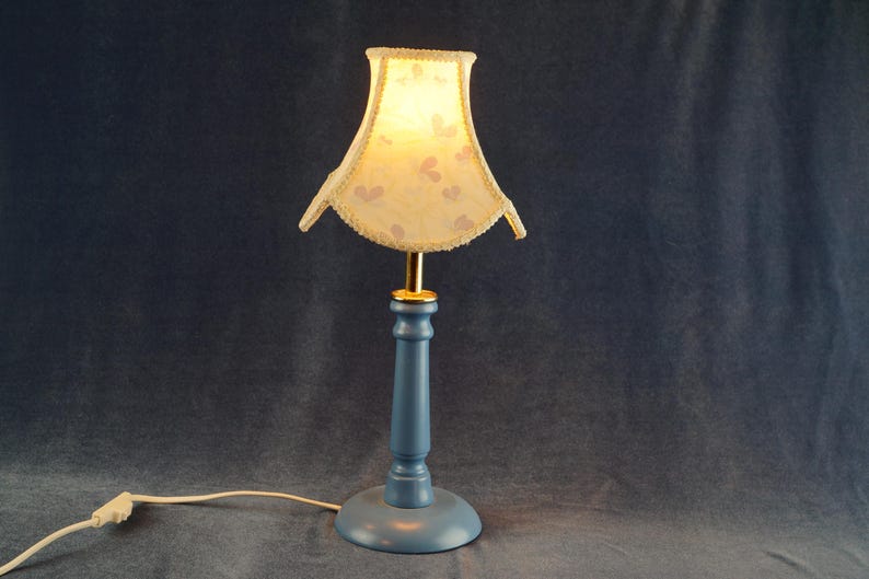 K&ouml;nnte beinhalten: Eine blaue Tischlampe mit einem wei&szlig;en und rosafarbenen Lampenschirm mit Muster. Der Lampenschirm hat einen gewellten Rand und eine goldene Verzierung. Der Lampenfu&szlig; ist einfach zylindrisch geformt.