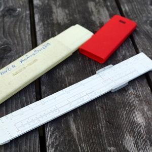 Puede incluir: Una regla de cálculo blanca con marcas negras y una funda roja. La regla de cálculo está en una funda blanca con el texto "T/IIHOR  Mancu-Orif" escrito en ella.