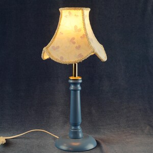 K&ouml;nnte beinhalten: Eine blaue Tischlampe mit einem wei&szlig;en Lampenschirm mit Blumenmuster. Der Lampenschirm hat einen goldenen Rand und der Lampenfu&szlig; ist aus Holz.
