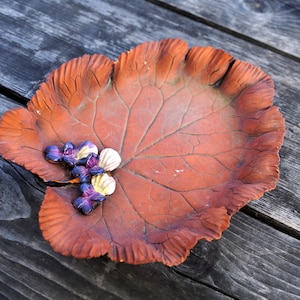 Op de afbeelding: Een terracotta schotel in de vorm van een blad met een getextureerd oppervlak en een verhoogde rand. De schotel is versierd met kleine paarse en witte bloemen.