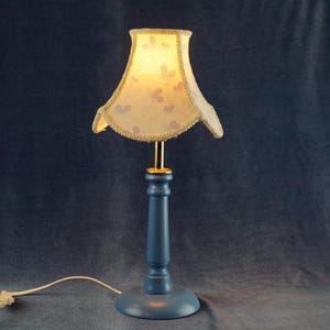 K&ouml;nnte beinhalten: Eine blaue Tischlampe mit einem wei&szlig;en Lampenschirm mit Blumenmuster. Der Lampenschirm hat einen gewellten Rand und einen goldenen Besatz.