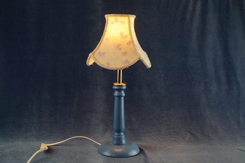 K&ouml;nnte beinhalten: Eine blaue Tischlampe mit einem wei&szlig;en Lampenschirm mit Blumenmuster. Der Lampenschirm hat einen gewellten Rand und einen goldenen Besatz.