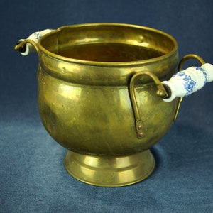 Può includere: Un vaso da fiori in ottone con corpo arrotondato e base larga. Ha due manici in porcellana con motivi floreali blu e bianchi.