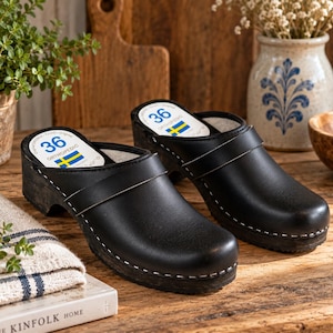 May include: A pair of black leather clogs with wooden soles and white stitching. The interior footbed features a white label with the number 36, the word ORTHOPEDIC, and a small Swedish flag icon. The clogs have a rounded toe and a wide strap.