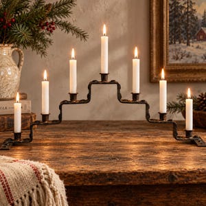 May include: A black metal candelabra with a stepped, tiered design holding seven white taper candles. The structure features a central elevated candle holder flanked by three holders on each side, all currently lit with glowing flames.