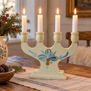 Könnte beinhalten: Ein cremefarbener Holzkandelaber mit vier Kerzenhaltern und brennenden weißen Stabkerzen. Der Sockel ist mit einem handgemalten blauen Blumenmotiv und goldenen Akzenten verziert. Die Struktur hat eine geschwungene Oberkante und einen rechteckigen Fuß.