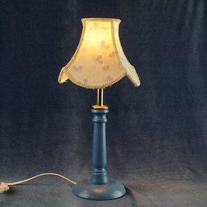 K&ouml;nnte beinhalten: Eine blaue Tischlampe mit einem wei&szlig;en Lampenschirm mit Blumenmuster. Der Lampenschirm hat einen gewellten Rand und eine goldene Einfassung.