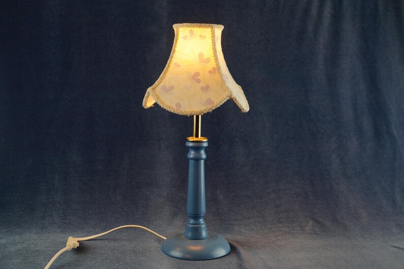 K&ouml;nnte beinhalten: Eine blaue Tischlampe mit einem wei&szlig;en Lampenschirm mit Blumenmuster. Der Lampenschirm hat einen gewellten Rand und eine goldene Einfassung.