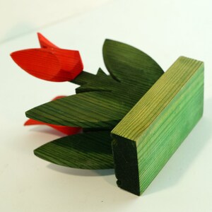 May include: A wooden sculpture of a flower arrangement with red tulips and green leaves. The sculpture is mounted on a green wooden base.