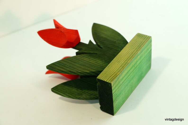 May include: A wooden sculpture of a flower arrangement with red tulips and green leaves. The sculpture is mounted on a green wooden base.