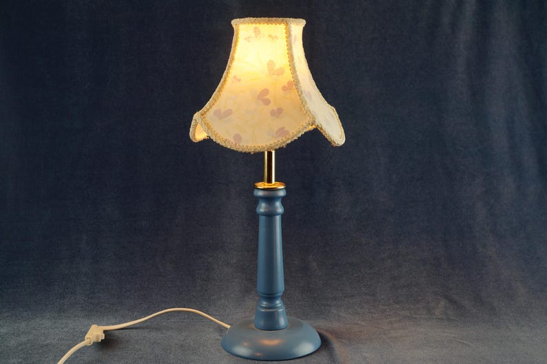 K&ouml;nnte beinhalten: Eine blaue Tischlampe mit einem wei&szlig;en Lampenschirm mit Blumenmuster. Der Lampenschirm hat einen goldenen Rand und der Lampenfu&szlig; ist aus Holz.