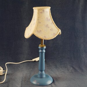 K&ouml;nnte beinhalten: Eine blaue Tischlampe mit einem wei&szlig;en Lampenschirm mit Blumenmuster. Die Lampe hat ein Kabel und einen Stecker.