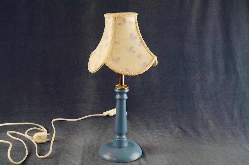 K&ouml;nnte beinhalten: Eine blaue Tischlampe mit einem wei&szlig;en Lampenschirm mit Blumenmuster. Die Lampe hat ein Kabel und einen Stecker.