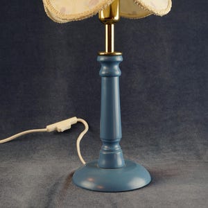 K&ouml;nnte beinhalten: Eine blaue Holztischlampe mit einem wei&szlig;en und goldenen Blumenlampenschirm. Die Lampe hat ein wei&szlig;es Kabel und einen Stecker.