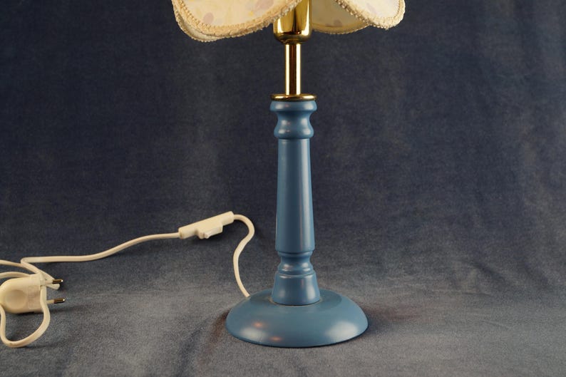 K&ouml;nnte beinhalten: Eine blaue Holztischlampe mit einem wei&szlig;en und goldenen Blumenlampenschirm. Die Lampe hat ein wei&szlig;es Kabel und einen Stecker.