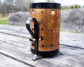 Puede incluir: Un portavelas de cobre con un asa y patas de metal negro. El portavelas tiene un diseño de copo de nieve y es perfecto para agregar un toque de encanto rústico a la decoración de tu hogar.