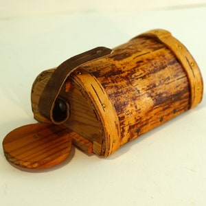 Pode incluir: Uma pequena caixa de madeira com uma alça de couro e detalhes em latão. A caixa é feita de madeira com um acabamento natural e tem uma tampa redonda.