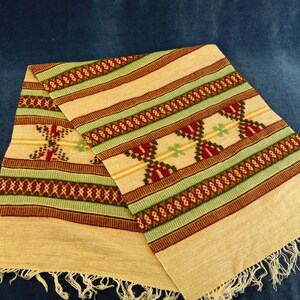 May include: A beige woven table runner with green, red, and brown geometric patterns. The runner has fringe on the ends.