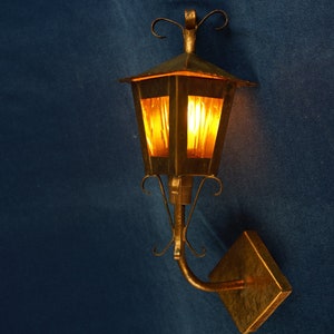 Op de afbeelding: Een vintage-stijl wandlamp met een gouden afwerking en heldere glazen panelen. De lamp heeft een decoratief scrollwerk ontwerp en wordt verlicht met een warme gloeilamp.