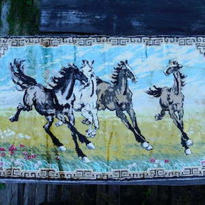 May include: A tapestry rug with a blue, green, and yellow background featuring four horses running. The horses are black, white, and brown. The rug has a fringed border with a geometric pattern.
