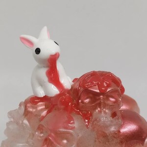 May include: A white rabbit figurine with pink ears and black eyes, appearing to bleed red liquid. The rabbit sits atop a cluster of red and clear skulls. The figurine has a glossy finish.