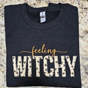 May include: Black crewneck sweatshirt with the words "feeling WITCHY" in gold and a patterned font. The letters in "WITCHY" have a beige and black pattern. The sweatshirt is made of a soft material.