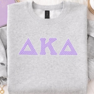 May include: A light gray sweatshirt with the Greek letters AKA in lavender with white polka dots. The text "Est. 1908" is below the letters. The sweatshirt is folded on a white surface.