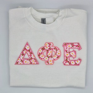 May include: White sweatshirt with a pink and white floral Greek letter design that reads "AΦE".