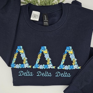 May include: Navy blue sweatshirt with embroidered floral Delta Delta Delta letters. The letters are decorated with blue, yellow, and white flowers. The words "Delta Delta Delta" are embroidered below the letters in a script font.