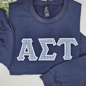 May include: Navy blue sweatshirt with the Greek letters AΣT in white and blue gingham fabric. The sweatshirt is folded, with the sleeves visible. A sprig of greenery is in the background.