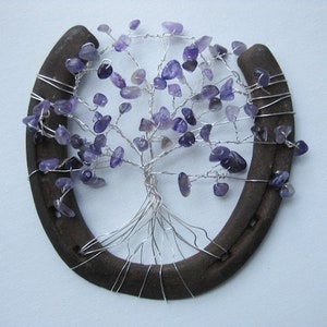 May include: A handcrafted tree of life sculpture made with silver wire and amethyst gemstones, set within a rustic horseshoe. The purple gemstones represent the leaves of the tree. The horseshoe is dark brown.