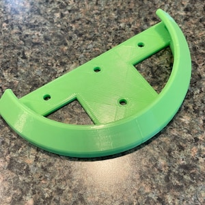 Puede incluir: Una pieza de plástico verde impresa en 3D con una forma curva y varios agujeros. La pieza está diseñada para ser unida a otro objeto.