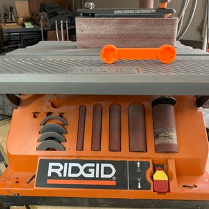 May include: An orange and gray Ridgid belt sander with a sanding belt and a variety of sanding drums. The sander has a built-in storage compartment for sanding accessories. The sander is on a wooden surface.