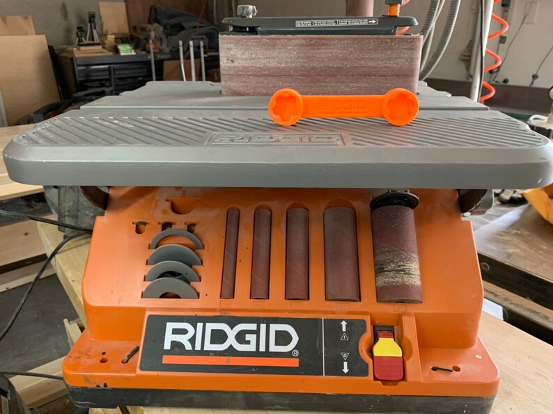 May include: An orange and gray Ridgid belt sander with a sanding belt and a variety of sanding drums. The sander has a built-in storage compartment for sanding accessories. The sander is on a wooden surface.