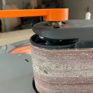 May include: Close-up of a belt sander with a black and orange handle. The sanding belt is brown and has a rough texture.
