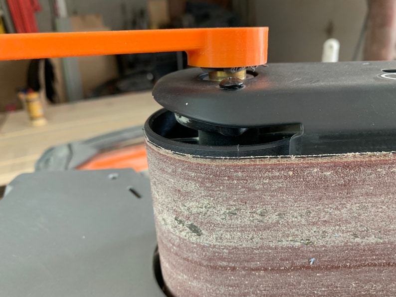 May include: Close-up of a belt sander with a black and orange handle. The sanding belt is brown and has a rough texture.