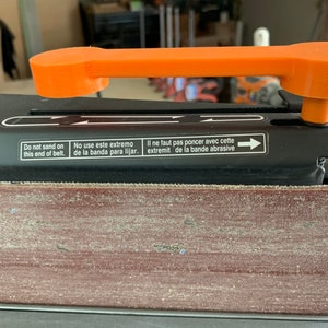 May include: Close-up of a black belt sander with an orange handle. The sander has a red sanding belt and a label that reads "Do not sand on this end of belt. No use este extremo de la banda para lijar. Il ne faut pas poncer avec cette extremit de la bande abrasive."