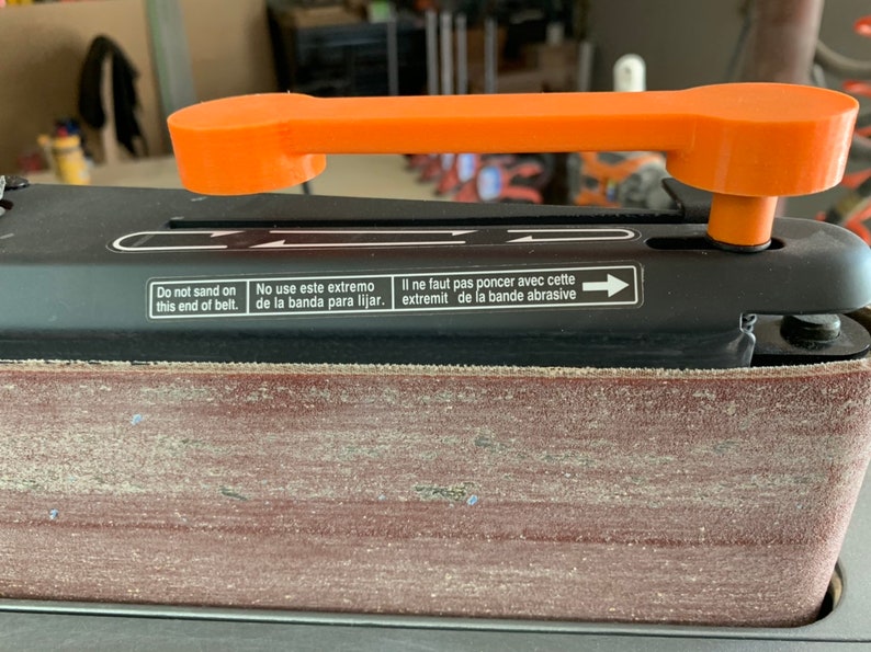 May include: Close-up of a black belt sander with an orange handle. The sander has a red sanding belt and a label that reads "Do not sand on this end of belt. No use este extremo de la banda para lijar. Il ne faut pas poncer avec cette extremit de la bande abrasive."