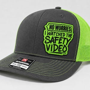 May include: A charcoal gray trucker hat with a neon green mesh back and matching neon green stitching on the brim. A patch on the front features the black text NO WORRIES I WATCHED THE SAFETY VIDEO on a neon green background with a black border.