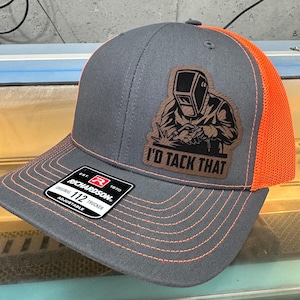 Könnte beinhalten: Eine graue und orangefarbene Trucker-Kappe mit einem braunen Leder-Patch, auf dem "I'd Tack That" steht. Der Patch zeigt eine Silhouette eines Schweißers. Die Kappe ist von Richardson 112.