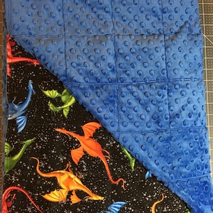 May include: A blue minky fabric blanket with a black background featuring a colorful dragon print. The blanket is folded in half, showing both sides.