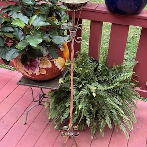 May include: A tall, slender metal candle stand featuring a twisted central pole, a circular top plate, and a three-legged base with decorative scrollwork. The stand has a weathered, metallic finish with copper and bronze tones throughout.