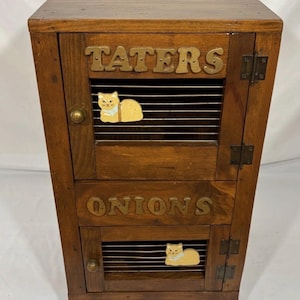 May include: A brown wooden storage bin with two stacked compartments. The top door features the word TATERS and a wire mesh panel with a yellow cat cutout. The bottom door features the word ONIONS and a wire mesh panel with a yellow cat cutout.