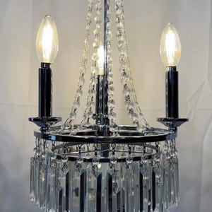 May include: A chrome chandelier with three light bulbs. The chandelier features hanging crystal accents and a circular design. The light bulbs are lit, and the fixture is suspended from a decorative top.