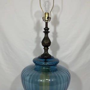 May include: A table lamp featuring a large, rounded, blue ribbed glass base. The lamp has a dark bronze-toned metal base with ornate detailing, a matching metal neck, and a brass-colored harp holding a single clear light bulb at the top.