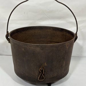 May include: A rustic, cast iron cauldron with a curved handle. The dark brown pot has three small legs and a decorative loop on the front. The cauldron is designed for cooking and is a vintage piece.