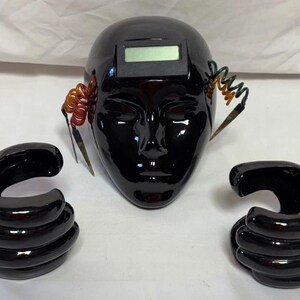 May include: A black ceramic sculpture of a face with a digital display on the forehead and two black ceramic hands.