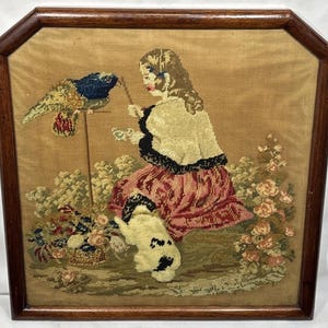 May include: A framed needlepoint tapestry featuring a person in a white shawl and red skirt sitting in a garden with a small dog, a bird on a perch, and floral accents. The artwork is enclosed in a dark brown wooden frame with angled top corners.