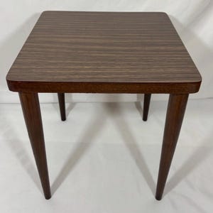 May include: A small, square side table with a dark brown, wood-grain top and four tapered legs. The table is approximately 20 inches tall and 18 inches wide, with rounded corners. The wood has a warm, natural finish.