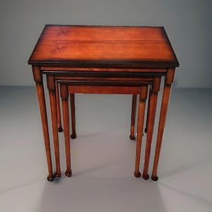 May include: A set of three nested tables featuring a rich, warm brown wood grain finish with darker, shaded edges. Each table has a rectangular top and four slender, straight legs that terminate in rounded, ball-shaped feet at the base.